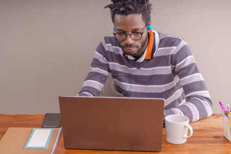 Téléchargez les photos : Étudiant afro-américain avec démarreur verrouille coiffure, profondément absorbé, regardant attentivement son écran d'ordinateur portable, assis à un bureau en bois avec écouteurs sans fil. - en image libre de droit