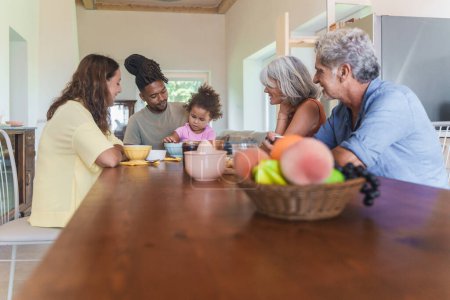 Photo for In a bright kitchen, a multiracial family gathers for breakfast. The grandparents, all attentively watch the biracial child as she picks cherries from a bowl, creating a heartwarming scene. - Royalty Free Image