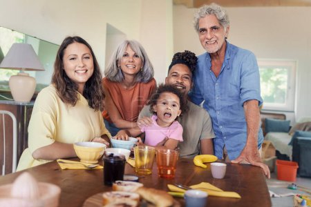 Photo for Cheerful extended family, including grandparents, parents, and a biracial child, pose together in a spacious, sunlit kitchen for a family portrait. - Royalty Free Image