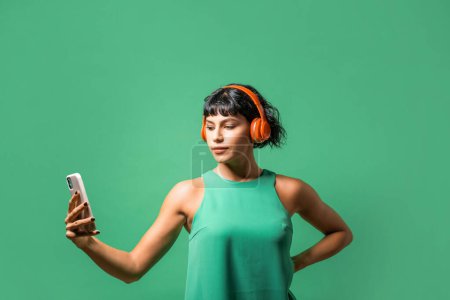 Téléchargez les photos : Portrait d'une jeune femme jouissant de musique sur son téléphone, portant un casque rouge et un débardeur vert sur un fond vert assorti. - en image libre de droit