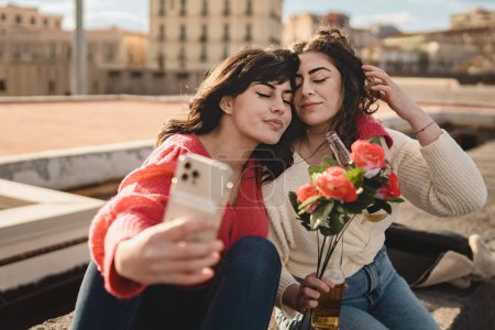 Foto de Una joven pareja de lesbianas de unos veinte años, tomando una selfie en un muelle, se acerca la cabeza con los ojos cerrados. Uno sostiene un ramo de rosas rojas. - Imagen libre de derechos