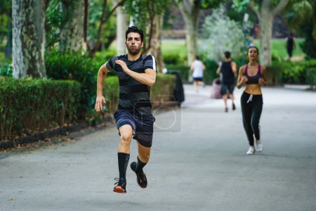 Foto de Adulto joven en ropa deportiva trotando en un sendero del parque, con compañeros corredores en el fondo debajo de los árboles. - Imagen libre de derechos