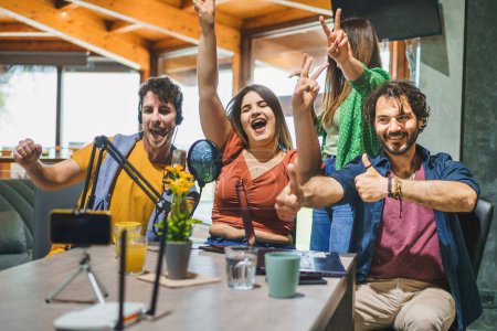 Photo for A group of four content creators jubilantly celebrate and gesture towards a smartphone mounted on a tripod, with a microphone in front of them, as they record a video stream for their video blog (vlog). - Royalty Free Image