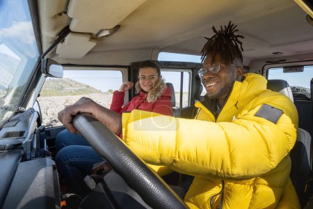 Téléchargez les photos : Homme africain en veste de randonneur conduit un véhicule hors route, avec sa compagne caucasienne le regardant avec un sourire chaleureux. - en image libre de droit