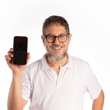 Foto de Un hombre alegre de cincuenta años de edad, con pelo rizado, barba y gafas, muestra con confianza una pantalla de teléfono inteligente, aislado en un fondo de estudio blanco. Lleva una camisa de polo y mira directamente a la cámara.. - Imagen libre de derechos