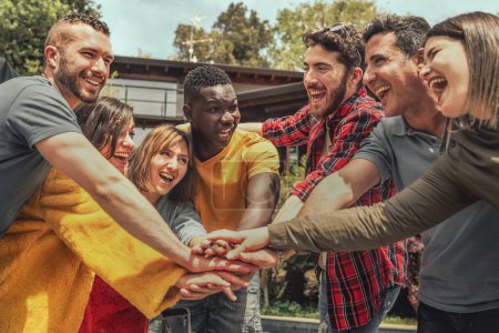 A multiracial group of young individuals forms a circle outdoors, placing their hands on top of one another's. They celebrate joyfully, looking into each other's eyes, embodying unity and friendship.