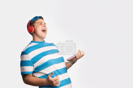 Foto de Un individuo no binario con llamativo cabello azul se pierde en la euforia de su canción favorita, con auriculares rojos vibrantes entregando los ritmos y un teléfono inteligente en la mano. - Imagen libre de derechos