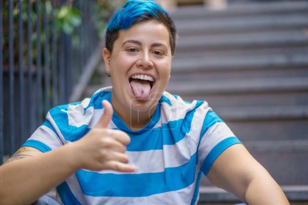 Photo for In a serene urban setting, a non-binary individual with vibrant blue hair exudes joy and confidence. With a cheeky tongue-out gesture, piercing, and a thumbs-up, they embrace the moment fully. - Royalty Free Image