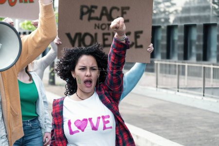 Téléchargez les photos : Une jeune femme fervente aux cheveux bouclés lève le poing d'un geste puissant, symbolisant la défiance et la demande de changement. Derrière elle, un autre manifestant tient un signe plaidant pour "LA PAIX PAS LA VIOLENCE" - en image libre de droit
