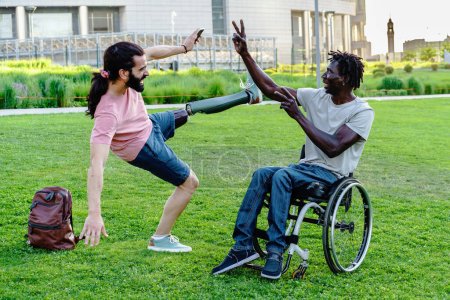 Foto de Dos amigos diversos al aire libre: Un hombre con una pierna protésica balancea juguetonamente, mientras que el otro, en una silla de ruedas, se extiende alegremente. Su vínculo irradia felicidad en medio de un espacio verde urbano. - Imagen libre de derechos