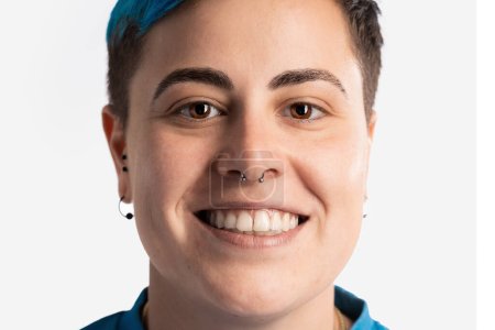Photo for Close-up portrait of a smiling gender fluid woman, showcasing her androgynous features. She has vibrant blue short hair, a nose piercing, and an earring, confidently displaying her teeth. - Royalty Free Image