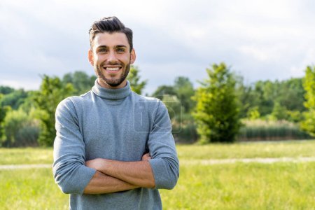 Photo for A handsome young Caucasian man with a beard stands confidently in an outdoor park, arms crossed. He offers a genuine smile, making direct eye contact with the camera. - Royalty Free Image