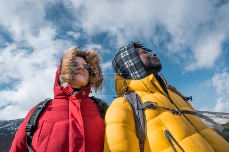 Photo for Two hikers, captured from a low angle against a cloudy blue sky, share a moment of wonder. The young man and woman, representing a blend of cultures, look upwards, symbolizing exploration and unity. - Royalty Free Image