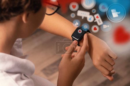 Photo for A boy seen from behind, uses his smart watch. Emoticon icons representing social network interactions emanate from the device, illustrating the digital world's connectivity and engagement. - Royalty Free Image