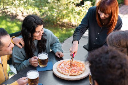 Téléchargez les photos : Une jeune femme découpe magistralement une pizza fraîchement cuite, tandis que des amis attendent avec impatience dans un cadre extérieur, tous prêts à plonger dans un repas détendu et joyeux ensemble. - en image libre de droit