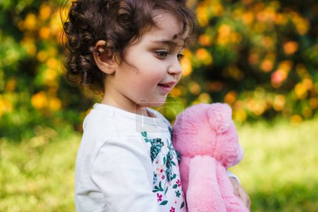 Photo for A curly-haired young girl clutches a pink plush toy, lost in thought amidst an autumnal park setting. The warm sunlight enhances the nostalgic and innocent ambiance of this genuine childhood moment. - Royalty Free Image