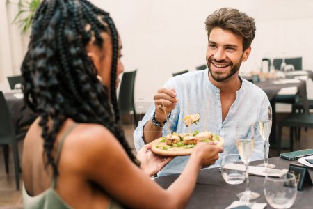 Téléchargez les photos : Un jeune homme souriant partage un repas avec une femme dans un cadre de restauration contemporain, soulignant un rendez-vous déjeuner détendu et agréable. Meilleurs amis appréciant la nourriture. - en image libre de droit