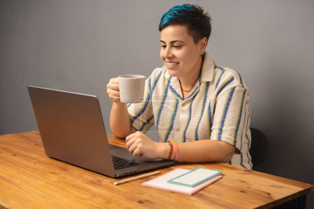 Téléchargez les photos : Une femme moderne et stylée aux cheveux bleus est assise à son bureau, sirotant du café et travaillant sur son ordinateur portable, mettant en valeur un cadre contemporain de travail à la maison.. - en image libre de droit