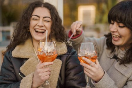 Téléchargez les photos : Deux femmes rayonnent de bonheur en dégustant leurs cocktails spritz à l'extérieur, capturant un moment spontané de joie et d'amitié. - en image libre de droit