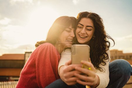 Téléchargez les photos : Deux femmes, éclatantes de rire, partagent un moment avec un smartphone. Le soleil se couche derrière, jetant une teinte dorée, résumant leur joie authentique et leur connexion profonde. - en image libre de droit