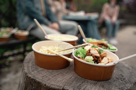 Photo for Bowls of noodles and tofu delicacies placed on a rustic wooden surface during an outdoor gathering. Blurred figures enjoy the meal, capturing a serene, communal moment. - Royalty Free Image