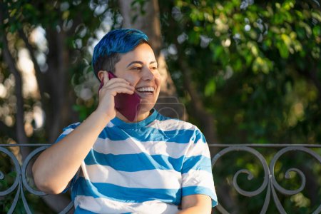 Photo for LGBTQ+ young woman with vibrant blue hair engaged in a cheerful phone conversation outdoors, seated against a backdrop of lush greenery, emanating a joyous and proud vibe. - Royalty Free Image
