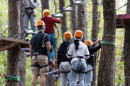 Téléchargez les photos : Un groupe se lance dans un défi de cordes hautes dans un parc d'aventure, mettant en valeur le travail d'équipe et l'agilité au milieu d'obstacles au sommet des arbres. - en image libre de droit