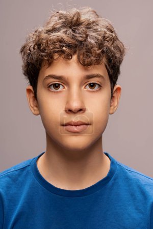Photo for A portrait of a young teen boy showcasing a pensive expression, curly hair prominently displayed, wearing a simple blue shirt against a neutral background. - Royalty Free Image