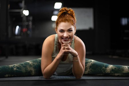 Foto de Una joven con una sonrisa radiante exhibe la alegría de mantenerse activa mientras se detiene entre las poses de yoga durante su entrenamiento en el gimnasio. - Imagen libre de derechos