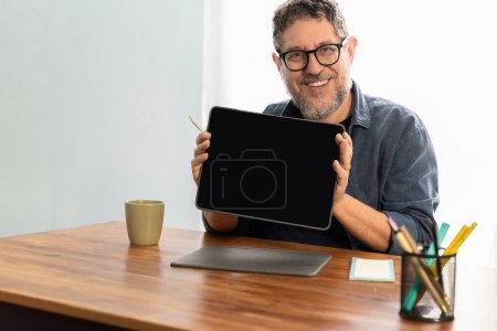 Foto de Un hombre alegre de cincuenta años que muestra con confianza la pantalla de su tablet digital mientras mira directamente a la cámara, encarnando la accesibilidad y el compromiso con la tecnología. - Imagen libre de derechos