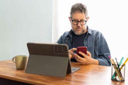 Photo for Man in a home office using a smartphone with a tablet in the background, showcasing efficient use of digital tools. - Royalty Free Image