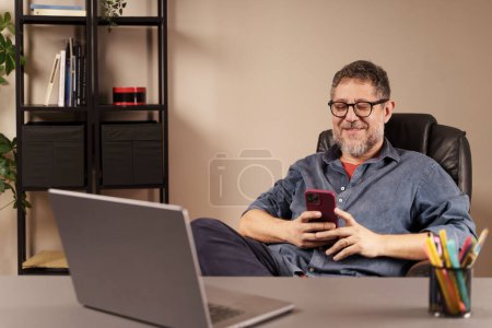 Téléchargez les photos : Homme d'âge moyen souriant à son smartphone, travaillant confortablement à partir d'un bureau à domicile bien organisé. - en image libre de droit