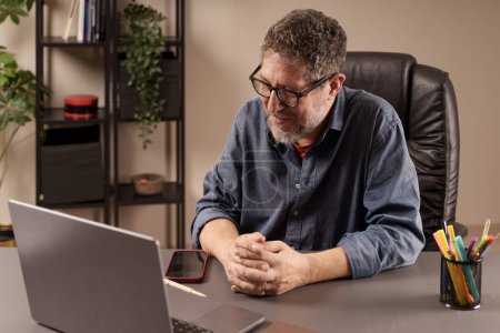 Hombre en la oficina del hogar sonriendo en la pantalla de su computadora portátil, posiblemente involucrado en una videollamada agradable o viendo contenido.