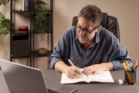 Photo for Man focused on writing in his planner, organizing tasks while working on his laptop in a home office setting. - Royalty Free Image