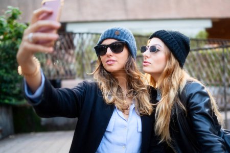 Photo for Two fashionable women wearing beanies and sunglasses taking a selfie on a sunny day, capturing a moment of friendship. - Royalty Free Image