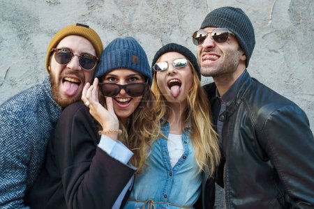 Photo for A joyful group of young adults wearing beanies and sunglasses making playful faces for a group selfie. - Royalty Free Image