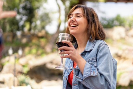 Téléchargez les photos : Une femme vêtue d'une veste en jean rit tout en dégustant un verre de vin rouge, capturant un moment de pure joie. - en image libre de droit