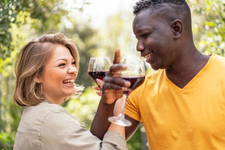 Téléchargez les photos : Un couple interracial partage un toast avec des verres de vin rouge, leur rire symbolisant un lien joyeux. - en image libre de droit