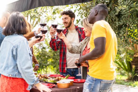 Téléchargez les photos : Un vibrant rassemblement en plein air avec divers amis griller avec des verres à vin, célébrer une occasion joyeuse avec un barbecue en arrière-plan. - en image libre de droit