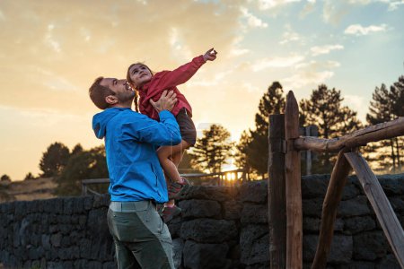 Vater hebt seine Tochter spielerisch in die Höhe, während sie gemeinsam einen Sonnenuntergang genießen, und fängt einen freudigen Moment familiärer Bindung ein.