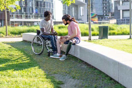 Téléchargez les photos : Discuter dans le parc entre deux hommes, mettant en valeur l'inclusivité avec l'un sur un fauteuil roulant et l'autre avec une jambe prothétique. - en image libre de droit