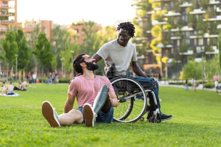 Foto de Dos hombres compartiendo risas en un parque, uno con una pierna protésica y el otro en una silla de ruedas, disfrutando de un ocio inclusivo al aire libre. - Imagen libre de derechos