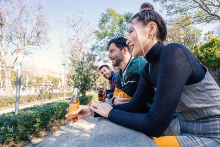 Téléchargez les photos : Un groupe diversifié de jeunes adultes rient et discutent tout en se reposant sur un mur du parc, partageant des boissons par une journée ensoleillée. - en image libre de droit