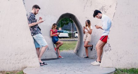 Foto de Cuatro adultos jóvenes, absortos en sus teléfonos inteligentes, se distinguen en un entorno urbano moderno, que simboliza la desconexión contemporánea en medio de la conectividad. - Imagen libre de derechos