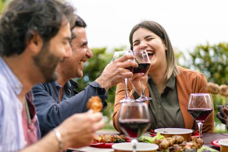 Photo for Joyful friends sharing a toast with red wine, laughing over a meal at an outdoor table surrounded by greenery - Royalty Free Image