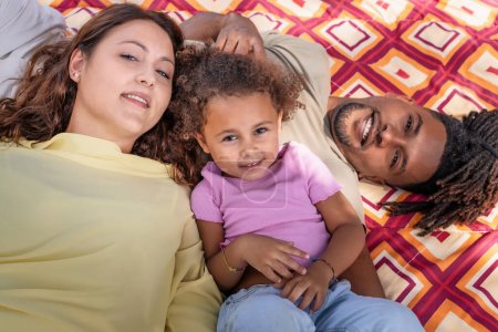 Téléchargez les photos : Une famille multiraciale avec un jeune enfant profite d'un moment de détente ensemble tout en s'allongeant sur une couverture à l'extérieur, mettant en valeur une scène de bonheur et de lien familial. - en image libre de droit