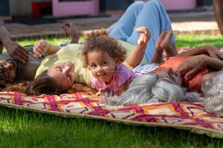 Foto de Familia Interracial comparte un picnic alegre, con un niño pequeño sonriendo brillantemente y mirando a la cámara, disfrutando de un cálido día de verano. - Imagen libre de derechos