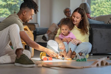 Photo for An interracial family immersed in play, with parents and child building a toy train track, under the watchful eyes of grandparents. - Royalty Free Image