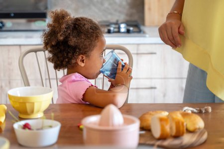 Téléchargez les photos : Le jeune enfant boit l'eau d'un verre, adoptant de saines habitudes à une table de cuisine à la maison, avec la famille à proximité. - en image libre de droit
