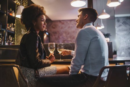 Foto de Sonriente pareja interracial disfrutando de un romántico brindis de vino en un acogedor bar, celebrando un momento especial juntos. - Imagen libre de derechos